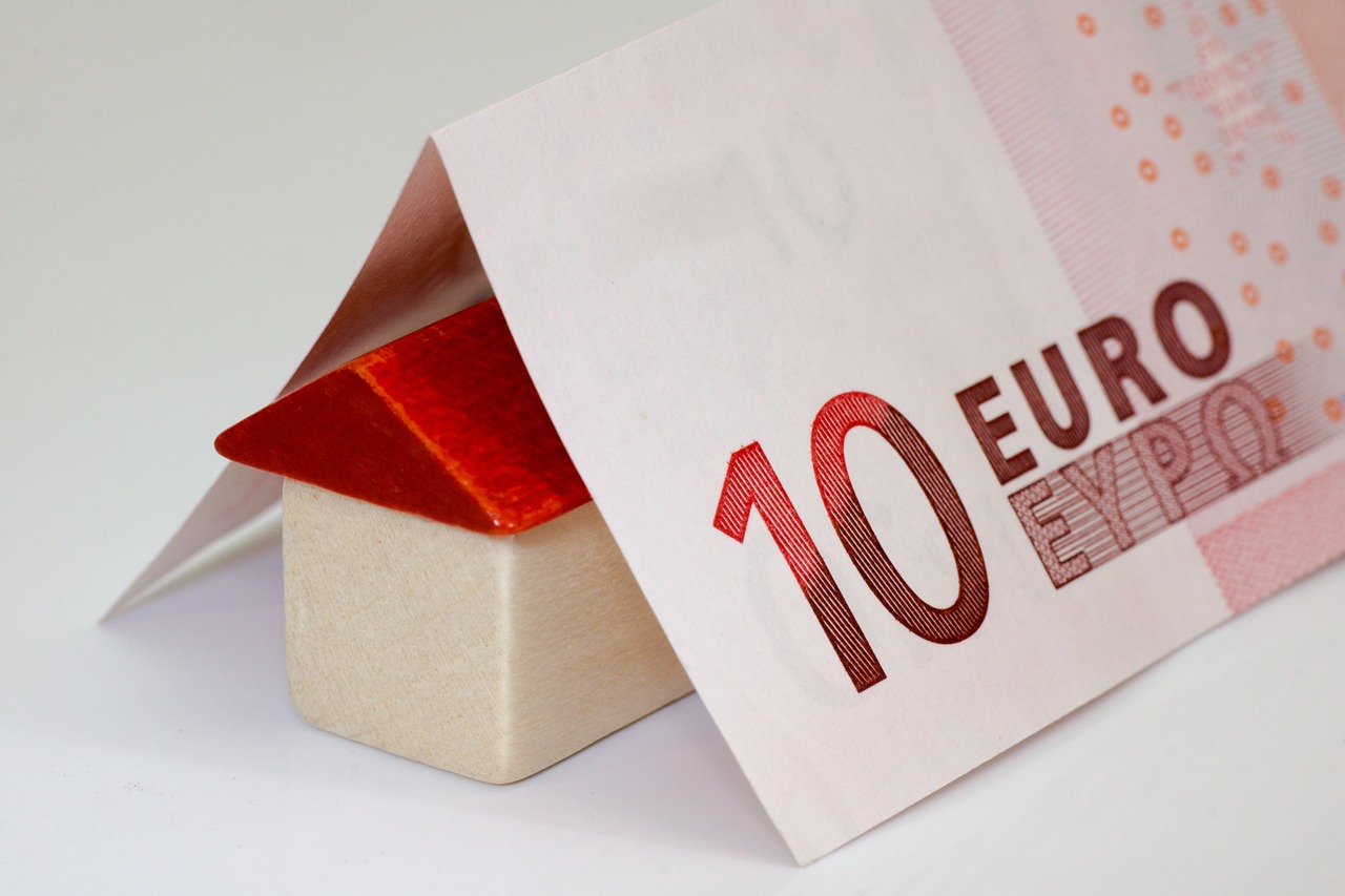 Kleines Holzhaus mit rotem Dach unter einem gefalteten Zehn-Euro-Schein als Symbol für die Kosten einer Immobilienfinanzierung